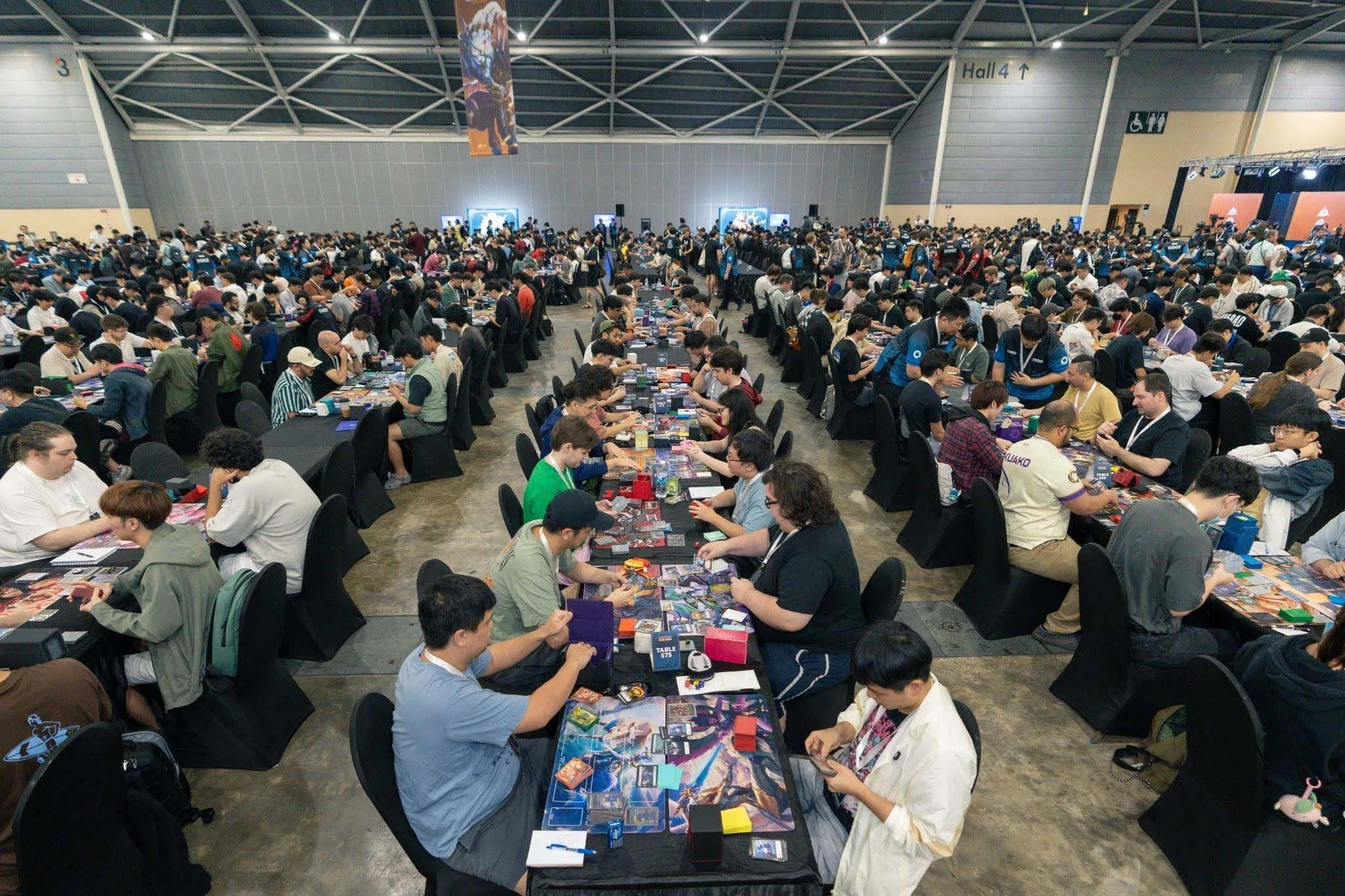 Riftbound competitive tournament hall Large hall filled with Riftbound trading card game competitors playing tournament matches at rows of tables.